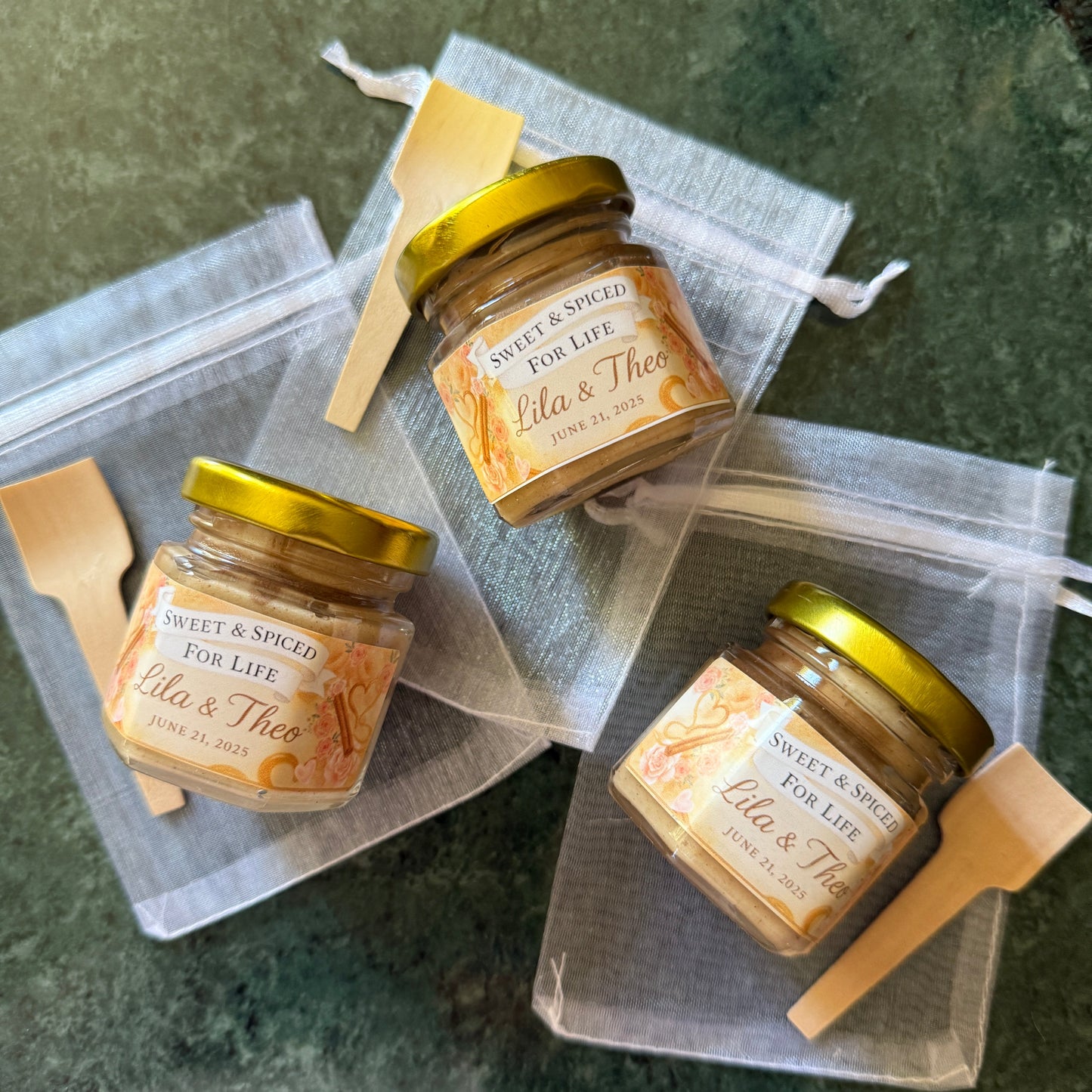 Flat lay of three creamed honey wedding favors in hexagon glass jars with personalized labels and soft blush tones